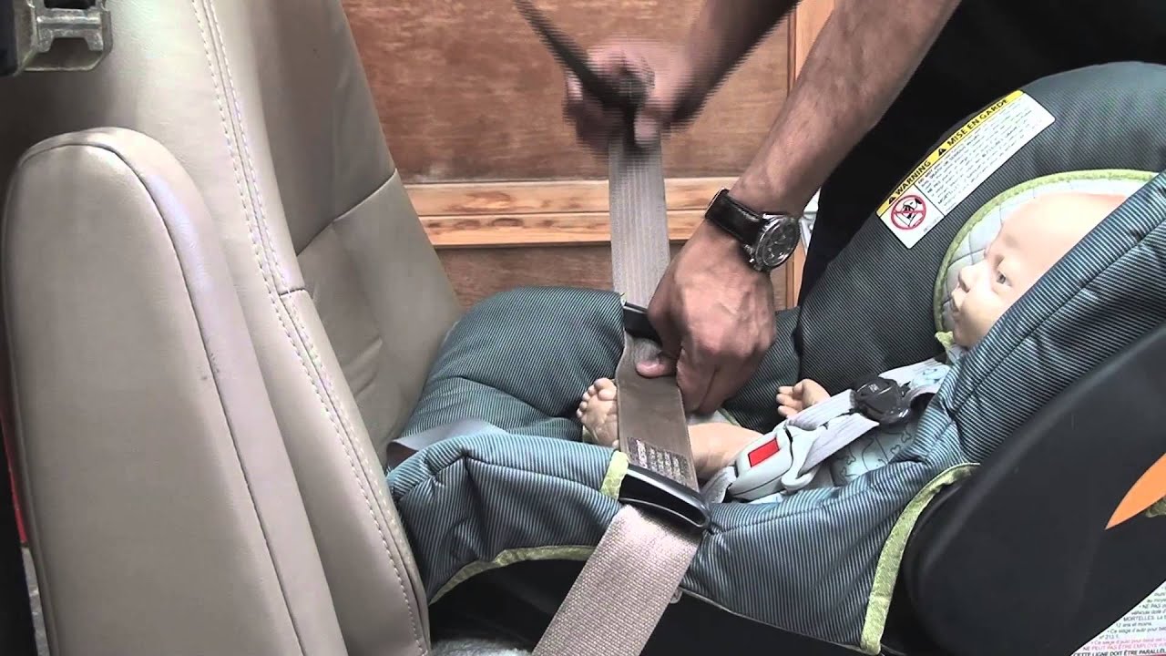 A parent positioning an infant car seat base on a used car's rear seat in a dealership parking lot, checking belt routing and angle indicator